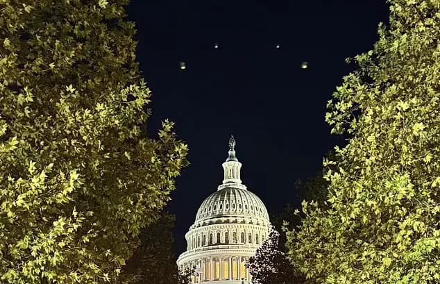 Lights in the night sky above the US Capitol, framed by trees